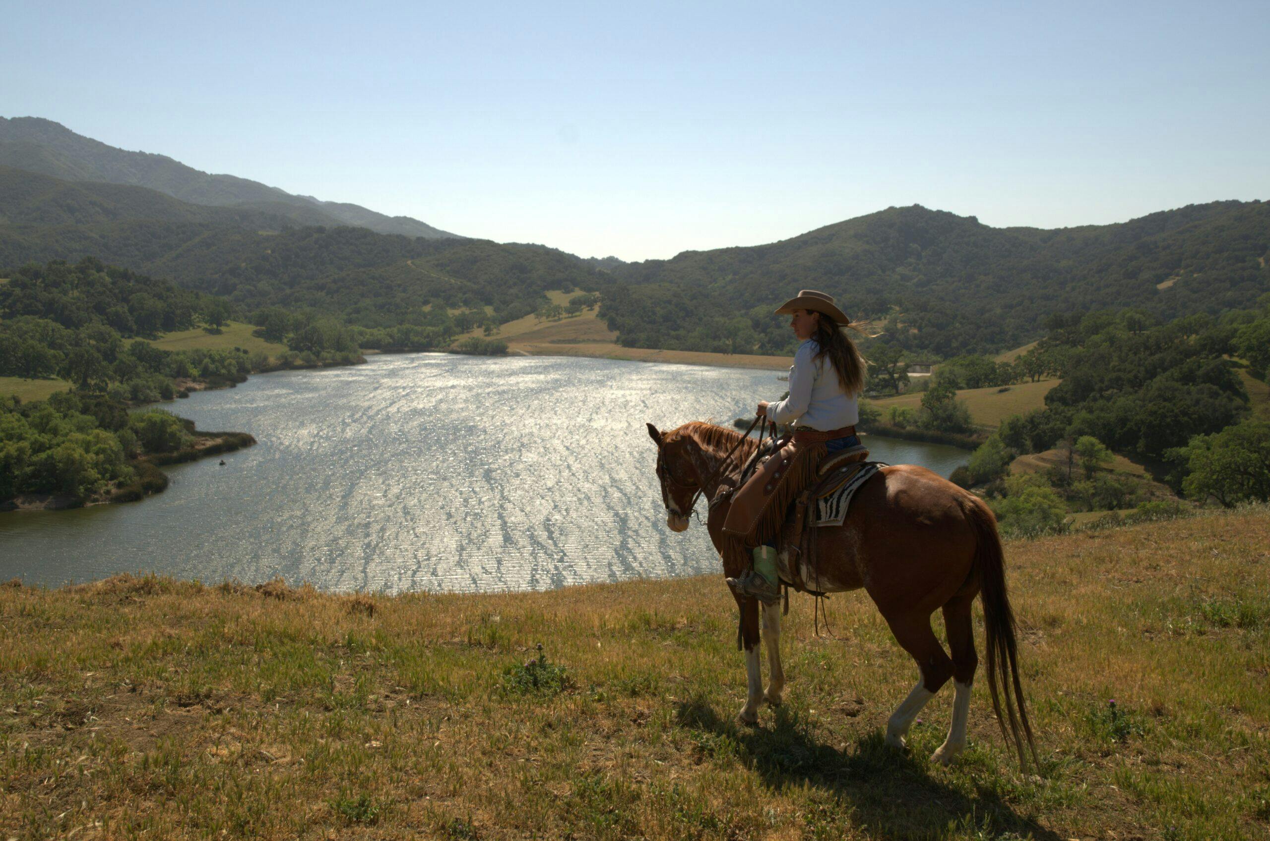 person riding horse on a grass hill overlooking the water