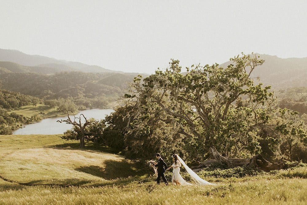 A bride & groom holding hands and running through a field of tall grass overlooking the lake at Alisal Guest Ranch & Resort